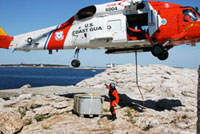 A Coast Guard helicopter hovers over a rock ledge on the ocean while a Coast Guard soldier maneuvers a large container having just been lowered down from the helicopter by a rope